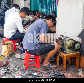 Three men sitting on plastic stools repairing shoes, working on the pavement on a street in Ho Chi Minh City, Vietnam. Stock Photo