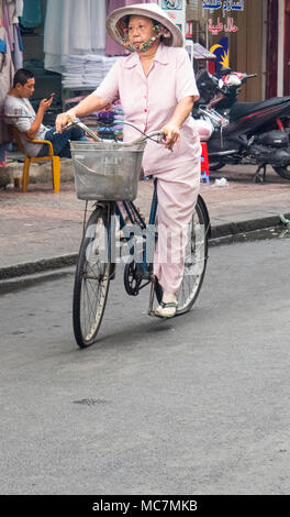 An elderly woman wearing a pink outfit and a conical hat cycling on a street in Ho Chi Minh City, Vietnam. Stock Photo