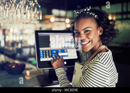 Smiling young African waitress wearing an apron using a touchscreen point of sale terminal while working in a trendy restaurant Stock Photo