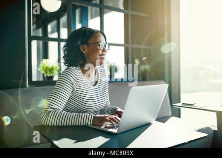 Smiling young African woman wearing glasses looking out of a window while sitting at a table working online with a laptop Stock Photo