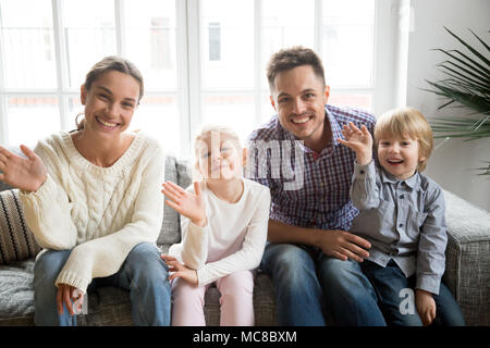 Portrait of happy multi-ethnic family with adopted kids waving hand looking at camera, smiling couple with children sitting on sofa making video call, Stock Photo