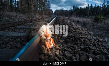 Train track with old doll lit by mystery light. Broken abandoned child toy at the rusty rail in forest. Idea of fate, apocalypse, sci-fi, war or abuse. Stock Photo