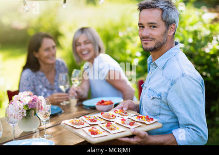group of friends in their forties gathered around a table in the garden to share a meal. A man offers snacks to guests while looking at the camera Stock Photo