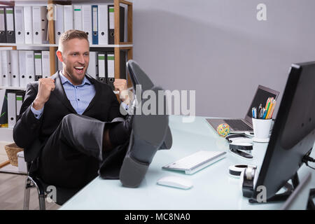Excited Businessman Looking At Computer With Feet On Desk In Office Stock Photo