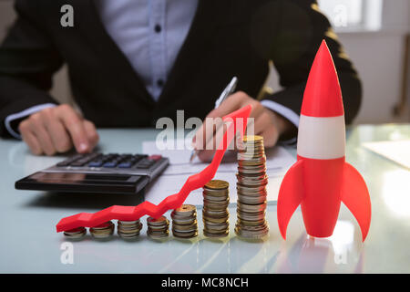 Close-up Of Red Rocket Besides Stacked Coins And Arrow Showing Upward Direction On Wooden Desk Stock Photo