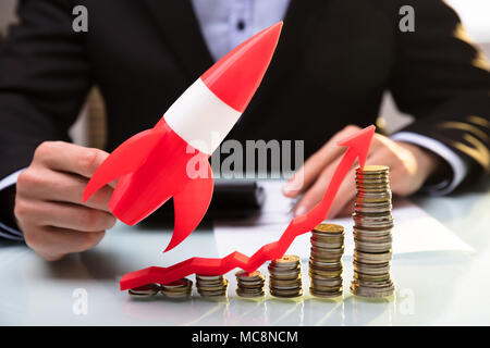 Businessperson's Hand Flying Red Rocket Over Stacked Coins And Arrow Showing Upward Direction Stock Photo