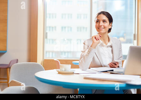 Inspired young businesswoman with smartphone and pen sitting by table and thinking of ideas while organizing work Stock Photo