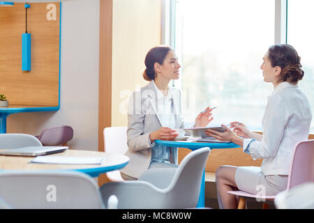 Two confident businesswomen sitting opposite one another by table in cafe and discussing working information for project Stock Photo