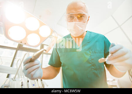 Mature dentist in uniform and protective mask and eyeglasses holding drill and mirror Stock Photo