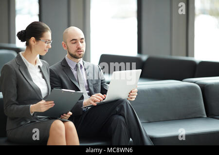 Two young delegates or agents sitting in airport lobby and discussing working moments while preparing for conference Stock Photo