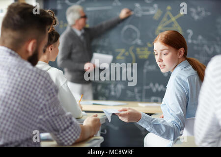 One of students passing note to her groupmate at lesson while teacher explaining formula on blackboard Stock Photo