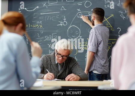 One of students writing with chalk on blackboard and explaining formula while teacher making notes in journal Stock Photo