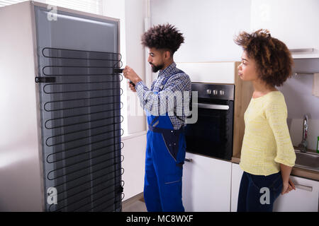 Woman Looking At Technician Repairing Refrigerator With Screwdriver In Kitchen Stock Photo