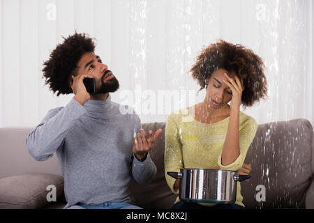 Young Man Calling Plumber While Woman Using Utensil For Collecting Water Leaking From Ceiling Stock Photo