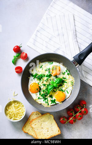 Fried eggs in pan, herbs, spices, cherry tomatoes, cheese toasts, basil for breakfast on grey concrete stone background. Copy space, top view, flat la Stock Photo