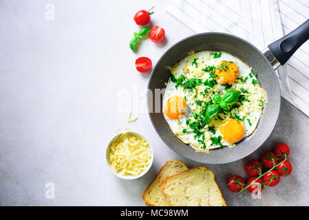 Fried eggs in pan, herbs, spices, cherry tomatoes, cheese toasts, basil for breakfast on grey concrete stone background. Copy space, top view, flat la Stock Photo