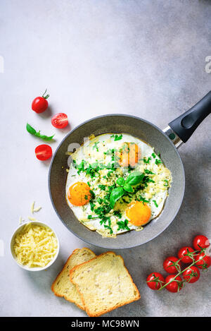 Fried eggs in pan, herbs, spices, cherry tomatoes, cheese toasts, basil for breakfast on grey concrete stone background. Copy space, top view, flat la Stock Photo