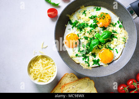 Fried eggs in pan, herbs, spices, cherry tomatoes, cheese toasts, basil for breakfast on grey concrete stone background. Copy space, top view, flat la Stock Photo