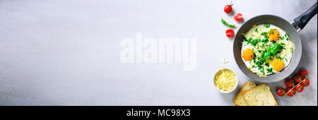 Fried eggs in pan, herbs, spices, cherry tomatoes, cheese toasts, basil for breakfast on grey concrete stone background. Copy space, top view, flat la Stock Photo