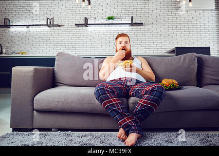 Thick funny man in pajamas eating a burger sitting on the couch, watching TV at home. Stock Photo