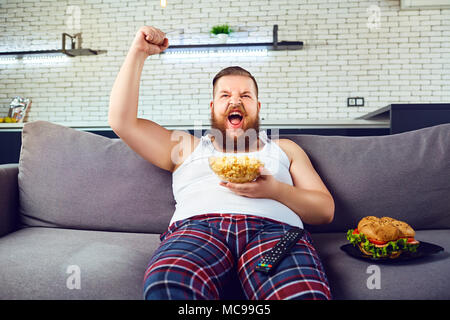 Thick funny man in pajamas eating a burger sitting on the couch, watching TV at home. Stock Photo