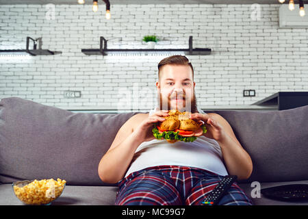 Fat funny man in pajamas eating a burger sitting on the sofa at home. Stock Photo