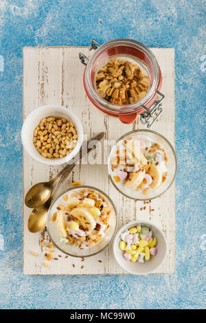 Healthy dessert from yogurt, banana, granola and nuts in glass glasses on a white wooden cutting board on a blue background. Top view Stock Photo