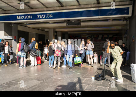 People walking and busker singer musician performing in front of the entrance of Brixton Underground station in South London, UK Stock Photo