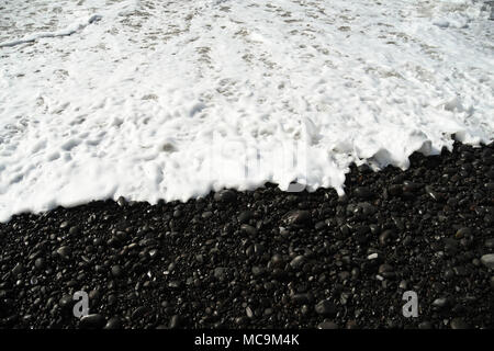 White foam from the incoming tide on the black volcanic beach Stock Photo