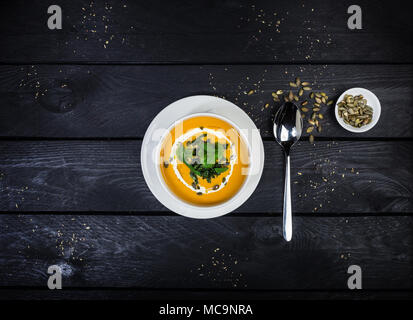 Pumpkin soup with jusai and fried cilantro, served in white bowl with a spoon, on the wooden background. Top view. Stock Photo