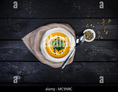 Pumpkin soup with jusai and fried cilantro, served in white bowl with a spoon and linen napkin, on the wooden background. Top view. Stock Photo