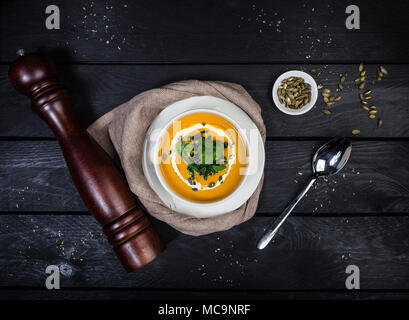 Pumpkin soup with jusai and fried cilantro, served in white bowl with a spoon linen napkin and pepper grinder on the wooden background. Top view. Stock Photo