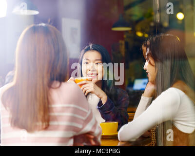 three happy beautiful young asian women sitting at table chatting talking in coffee shop or tea house, shot through window glass. Stock Photo