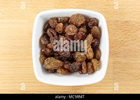 Overhead shot of raw sultanas, in a white bowl on a wooden chopping board background. Sultanas are dried seedless pale green grapes. Stock Photo