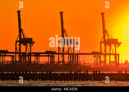 Orange red burnt sky at sunset against setting hot sun contrasting tall heavy industrial cargo loading cranes in Port Melbourne over Port Phillip bay  Stock Photo