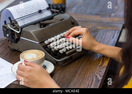 female hands holding a cup of cappucino and typing on retro typewriter placed on a wooden tabletop. Horizontal composition. Stock Photo