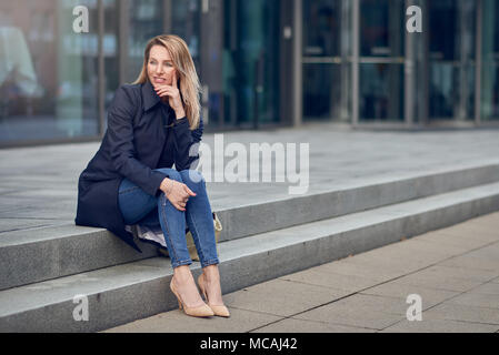 Attractive stylish woman in high heels and an overcoat sitting on urban steps looking thoughtfully to the side with her chin on her hand Stock Photo