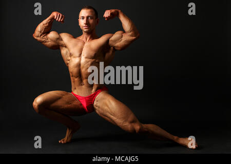 young male bodybuilder standing knee bend one leg squat showing front double biceps pose studio photo with black background mcajg5