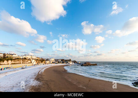 Beach partly covered in snow at Broadstairs resort. Landscape view, town and harbour with blue sky and white clouds. Lone man walking along seashore. Stock Photo