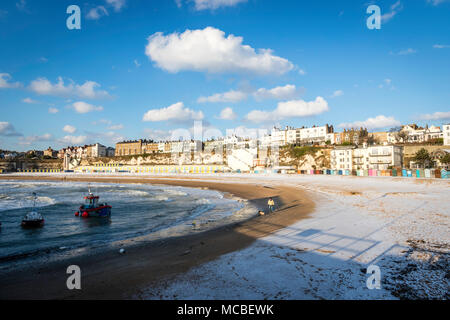 Viking bay beach at Broadstairs after snowfall in winter. Man walking dog along sea's edge. Town in background. Daytime, sunshine, blue sky. Stock Photo