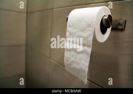 Close-up of white soft tissue paper roll in bathroom, restroom, wc on light brown beige tiling background ion simple shiny chrome holder. Modern lifes Stock Photo