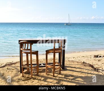 Romantic seascape with wooden table and chairs on sandy beach and a sailboat in the distance. Stock Photo