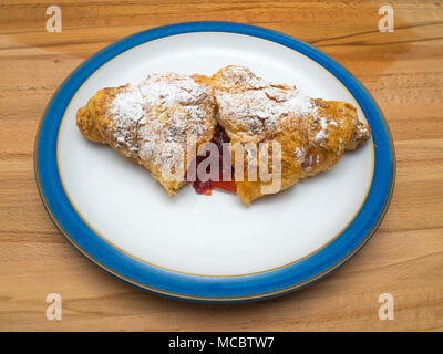 A sugared croissant split open to show Strawberry jam inside on a blue rimmed plate on a wooden table Stock Photo