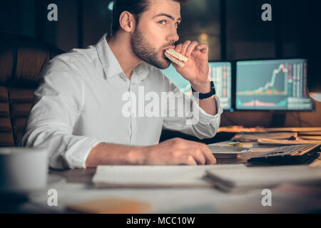 Busy businessman having late lunch eating sandwich in office working at night, stock broker trading online and eating snack looking at computer, workp Stock Photo
