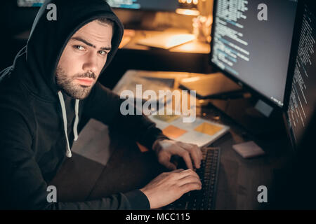 Dangerous hooded hacker looking at camera coding on computer organizing cyber ddos attack, programmer in hood hacking spying or stealing secret inform Stock Photo