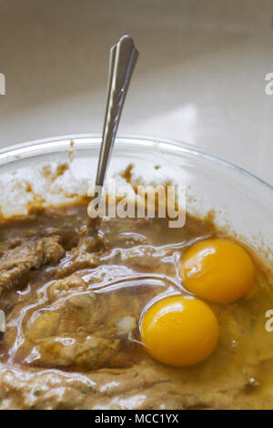 Close up of two raw, broken eggs lying on top of zucchini bread batter in a glass bowl with a spoon sticking out of it Stock Photo