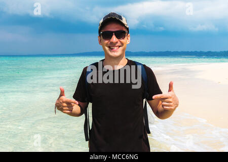 Casual man pointing with both hands to his blank gdark brown t-shirt on the background of beautiful exotic landscape with white sand beach and clear s Stock Photo