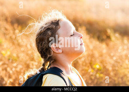 Summer girl with rucksack on sunset meadow smelling fresh air. Stock Photo