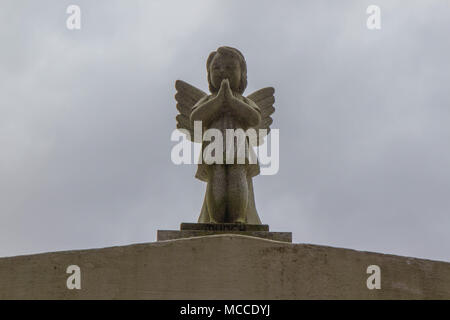 Stone sculpture of an angel on top of a tomb in Lafayette Cemetery #1 in New Orleans Louisiana, USA. Angel is a cherub in prayer, horizontal, gray sky Stock Photo