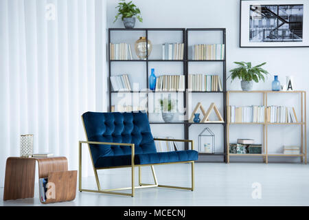 Navy blue armchair next to wooden table in cozy living room interior with bookshelf and poster Stock Photo